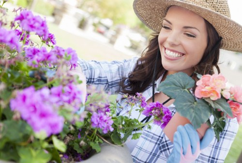 A young woman with a wide smile wearing a straw hat, light blue gardening gloves, and a checked shirt is tending to a variety of blooming garden plants in an outdoor space. The garden features a mix of vibrant purple and pink flowers, with some green foliage visible. The background shows a neatly maintained lawn area and some blurred trees or shrubs, indicating a well-kept garden environment typical of residential outdoor spaces in Sutton. The lighting suggests a bright, sunny day, enhancing the natural colours of the plants. This scene reflects garden maintenance and planting activities that Lawn Mowing Sutton offers for local garden care and landscaping services.